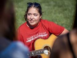 Gabriella Surace, a sophomore Music Therapy major at the John J. Cali School of Music, plays MIrrorball by Taylor Swift during a class held on the Student Center Quad. Alt Txt: A student holds a guitar while seated in the grass.